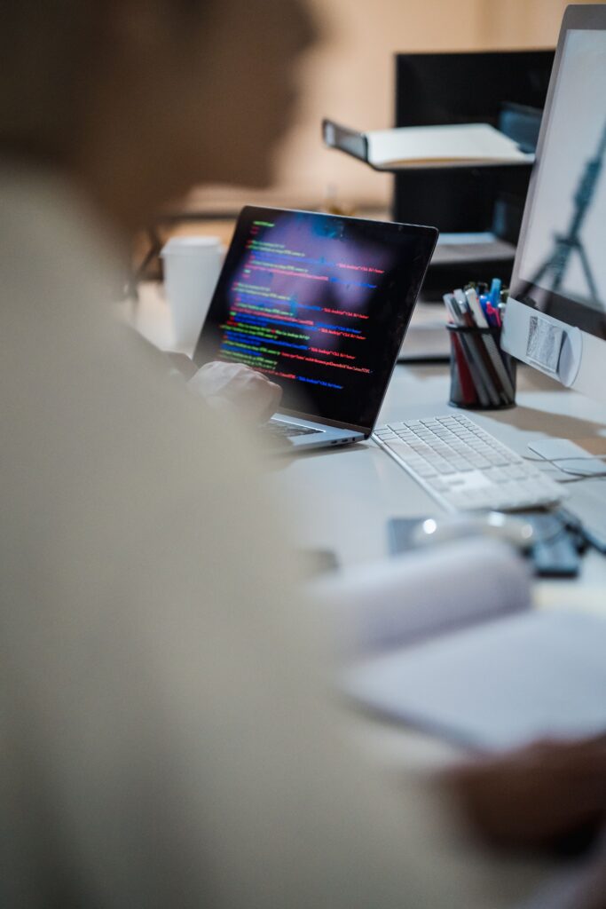 pexels photo 12899149 12899149 Blurred figure at a desk with a laptop displaying colorful code, indicating a programming environment.