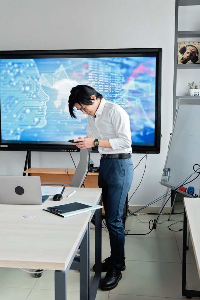 pexels photo 7988792 7988792 A man standing in an office checks his smartphone with a digital screen displaying AI graphics.